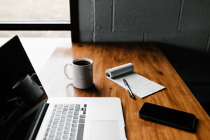 A laptop, cell phone, paper pad, and coffee mug on a wooden desk